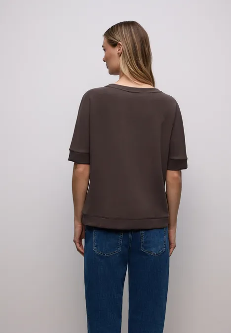 Frau von hinten trägt dunkles Kurzarmshirt und blaue Jeans vor weißem Hintergrund.